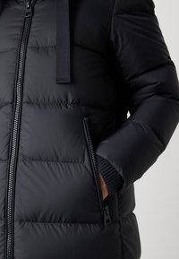 Zwarte pufferjack met een gewatteerde textuur, voorzien van een ritssluiting aan de voorkant en zijzakken. De mouwen zijn ribbelachtig voor extra warmte.