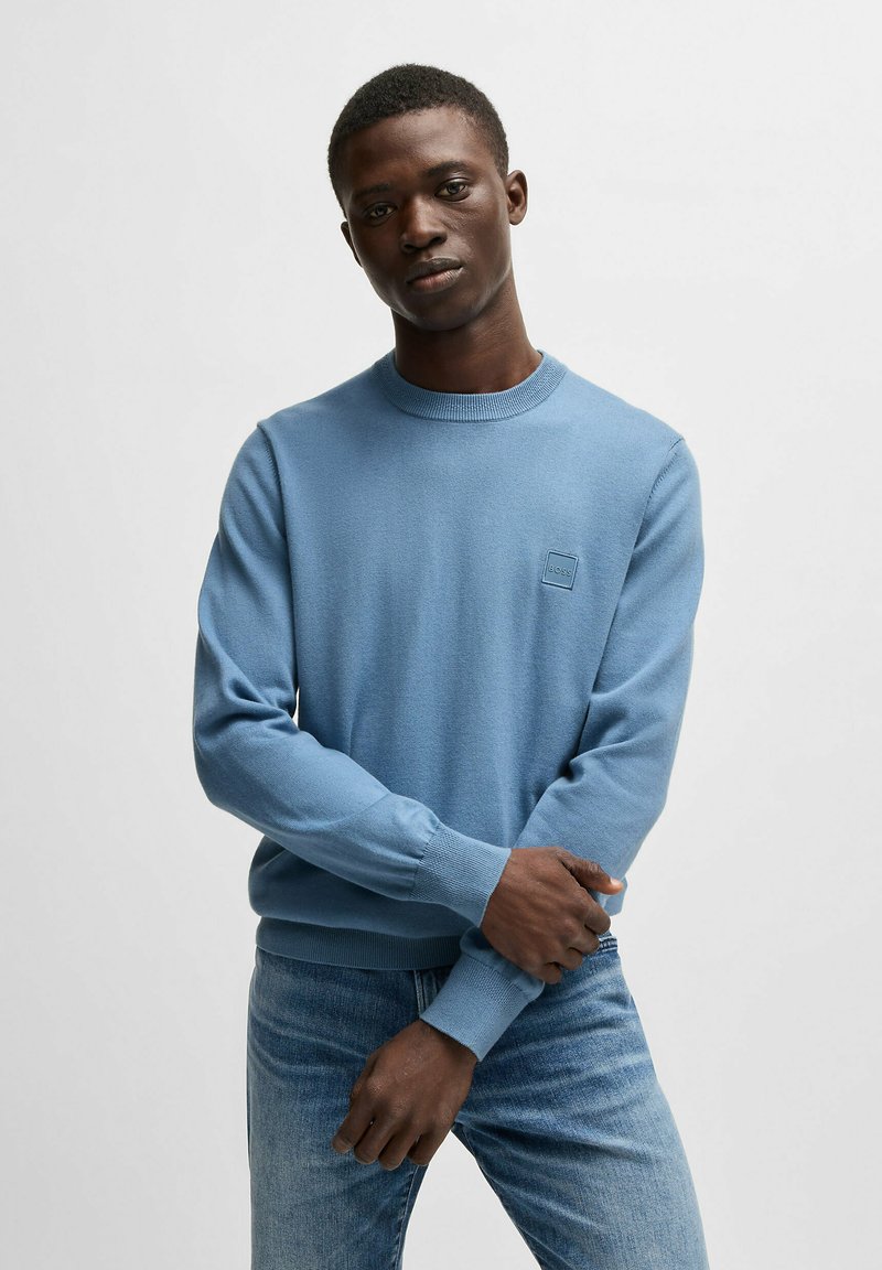 Hellblauer Rundhals-Pullover aus weichem Stoff, mit langen Ärmeln und gerippten Bündchen, sowie einem kleinen Logopatch auf der Brust.