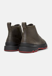 Stivali alla caviglia in pelle marrone con suole robuste nere e rosse, linguette posteriori e zip laterali, mostrati da dietro su uno sfondo bianco.