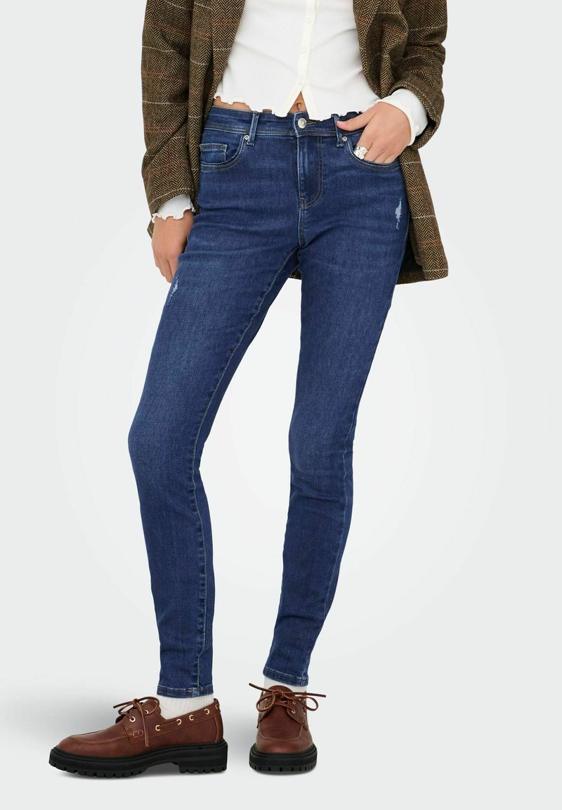 Kitsad sinised teksapüksid keskmise kõrgusega vöökoht, detailid ees olevates taskutes ja kulunud aktsendid. Kombineeritud pruunide nööriga saapadega.