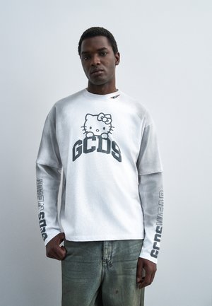 Čovjek nosi bijelu košulju dugih rukava s grafikom Hello Kitty i natpisom „GCDS“, uparenu s izblijedjelim zelenim trapericama na jednostavnoj svijetloj pozadini.