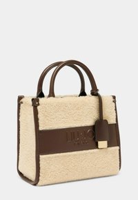 Sac à main marron et crème avec une texture en peluche et des accents en cuir lisse. Il dispose de poignées arrondies et d'une étiquette décorative. Nom de la marque embossé.