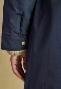 Giacca impermeabile blu navy con colletto in maglia texturizzata. Presenta un bottone a pressione dorato sul polsino. Vestibilità slim e tessuto liscio.