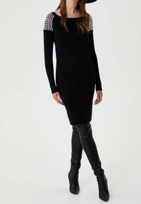 Robe noire en tricot avec un col bardot et des perles ornant les manches. Portée avec des bottes noires montantes jusqu'aux cuisses.