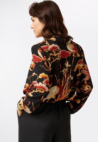 Schwarze Bluse mit buntem Blumen- und Vogelmotiv, ausgestattet mit einem Kragen, langen Ärmeln und einem lockeren Schnitt. Weicher Stoff mit glatter Textur.