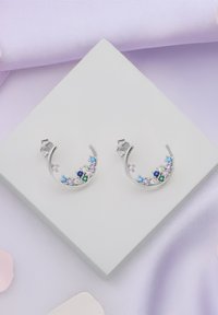 Silvriga hängörhängen med färgglada stenar i olika former och storlekar ordnade i en halvmåne-design, upplagda på en vit fyrkantig låda.