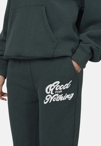 Zestaw ciemnozielonego hoodie i spodni dresowych. Hoodie posiada kieszeń z przodu, a na nogawce spodni znajduje się biały napis "Good for Nothing".