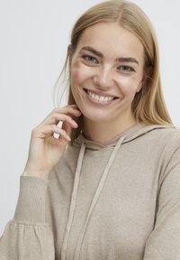 Hoodie beige avec une texture douce, capuche à cordon et poignets côtelés ajustés. Un design décontracté accentué par de douces manches bouffantes.