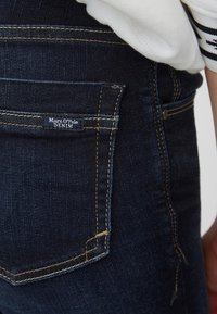 Los vaqueros de mezclilla de color azul oscuro presentan un bolsillo trasero con un acabado texturizado y una pequeña etiqueta que dice "Marc O'Polo DENIM." Costuras en contraste visibles.