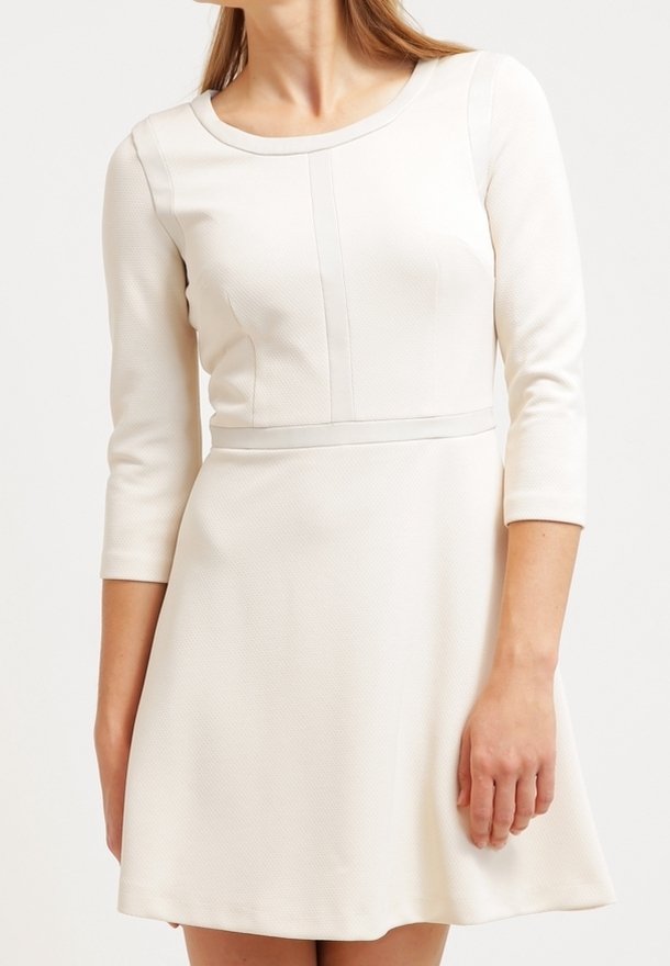 Femme portant une robe ajustée, blanche, texturée avec des manches trois-quarts et un col rond, debout devant un fond uni.