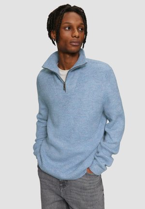 Pull bleu clair en maille côtelée avec un col zippé à moitié. Texture douce, coupe décontractée et manches longues. Porté sur un T-shirt blanc et un jean gris.