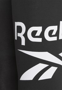 Fekete pamut póló, nagy fehér "Reebok" logóval és figyelemfelkeltő grafikával, éles vonalakkal. Simaság, kerek nyakú kivitel.