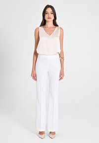 Blusa senza maniche di seta color crema con scollo a V, abbinata a pantaloni bianchi a vita alta e gamba larga e décolleté beige a punta. Bracciale d'oro come accessorio.
