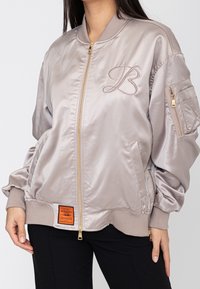 Veste bomber en satin taupe clair avec zip doré, "B" brodé sur la poitrine, poche zippée sur la manche et étiquette orange près de l'ourlet, portée par une personne.