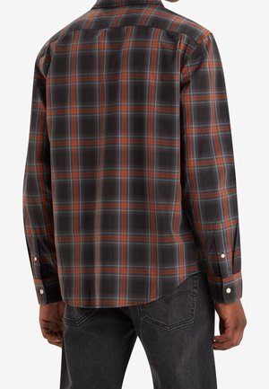 Mann trägt ein langärmeliges schwarz-orange-graues Karohemd, von hinten gesehen, kombiniert mit dunkelgrauen Jeans.