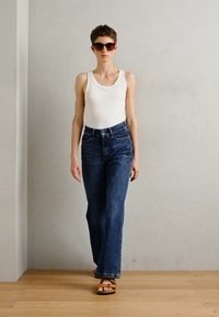 Jeans bleus taille haute, débardeur blanc, lunettes de soleil marron et sandales marron. Le mannequin marche sur un sol en bois avec un mur neutre en arrière-plan.