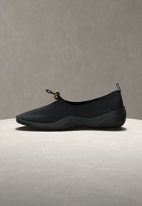 Μαύρο slip-on παπούτσι με στρογγυλεμένη μύτη, παχιά σόλα, ρυθμιζόμενο κουμπί επάνω και ταινία έλξης στη φτέρνα, πάνω σε ουδέτερη επιφάνεια.