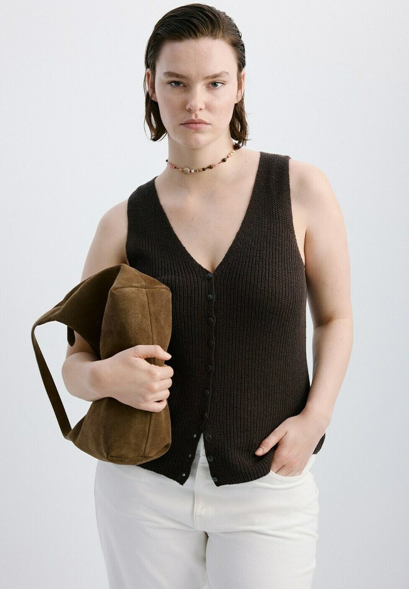 Femme portant un haut noir sans manches à boutons en tricot, un pantalon blanc, un collier de perles, tenant un sac à main en daim marron avec une main dans la poche.