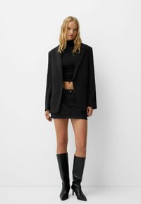 Blazer noir oversize sur un crop top noir, associé à une mini-jupe en denim noir et des bottes à talons aiguilles noires montantes.