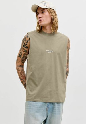 Mężczyzna z długimi włosami i tatuażami, ubrany w beżową koszulkę bez rękawów z logo "JJ Studios" oraz jasnoniebieskie dżinsy, patrzy w prawo, na głowie biała czapka.