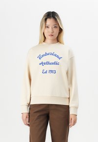 Hanorac crem cu text albastru "Timberland Authentic Est 1973." Dispune de un guler rotund și de manșete ribbate, având o croială lejeră.