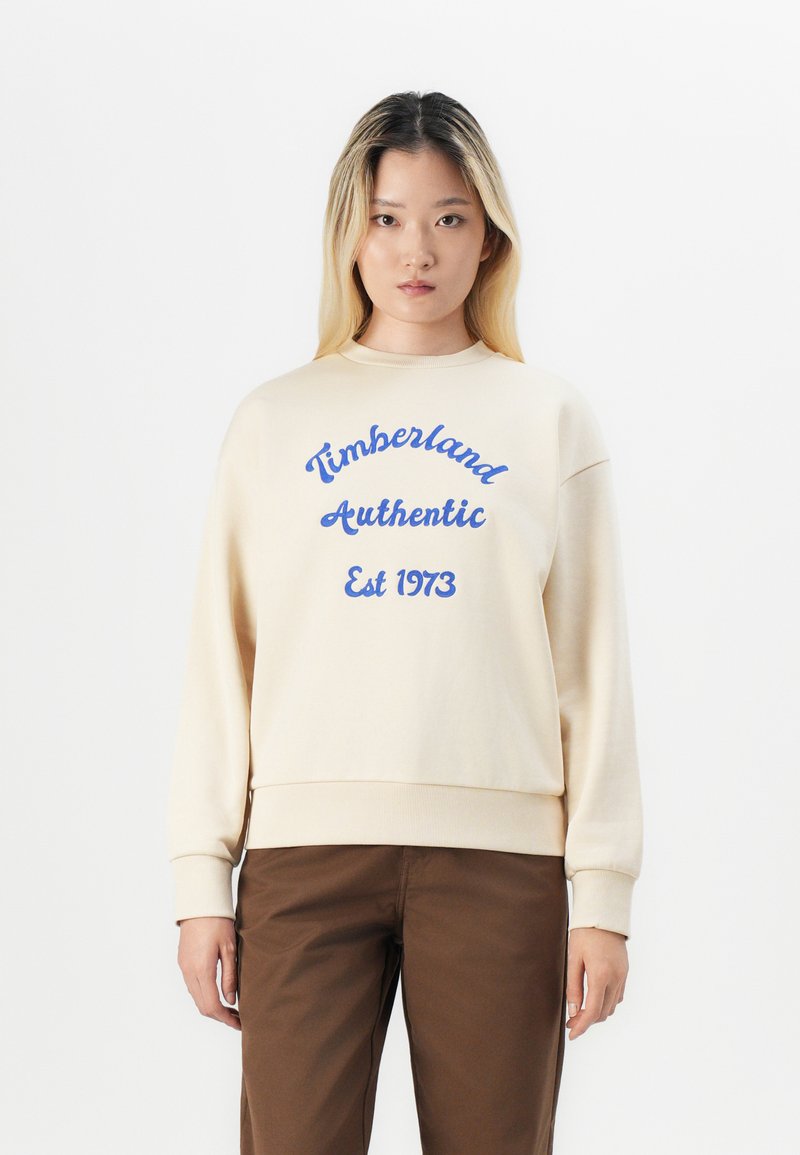 Hanorac crem cu text albastru "Timberland Authentic Est 1973." Dispune de un guler rotund și de manșete ribbate, având o croială lejeră.