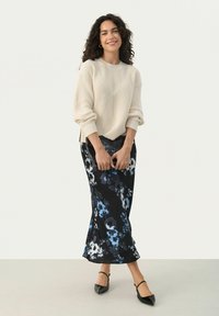 Crème ribbeltrui gecombineerd met een zwarte maxi-jurk met bloemenprint en blauwe en witte accenten. Het model houdt een zwarte clutch vast.