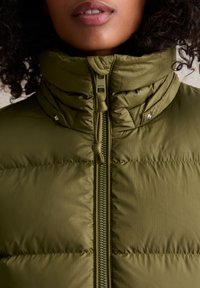Oliwkowa kurtka puchowa z wysokim kołnierzem, zamkiem błyskawicznym i ściągaczem. Posiada pikowaną fakturę i błyszczące akcenty z metalu.