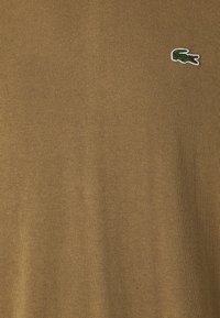 Pull en tricot marron avec un petit logo crocodile Lacoste sur la poitrine gauche. Le matériau est lisse avec une texture uniforme.