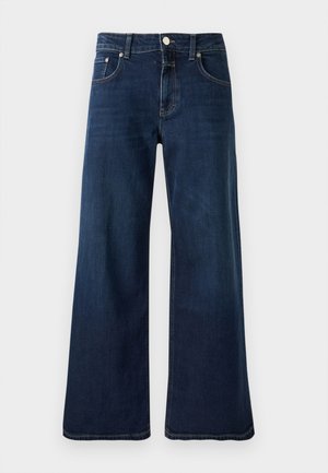 Donna in piedi che indossa un cardigan rosa chiaro, dolcevita crema, jeans a gamba larga blu scuro e stivaletti marroni su uno sfondo semplice.
