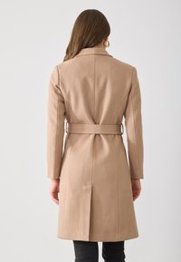Cappotto di lana beige chiaro con vita aderente, maniche lunghe, spacco posteriore e cintura, mostrato da dietro su uno sfondo neutro.