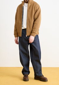 Brun ribbad jacka med dragkedja, vit t-shirt, lösa mörka denimbyxor och bruna skor med tjock sula. Enkel, funktionell design.