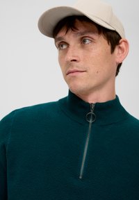 Tee-farbener Strickpullover mit Reißverschluss, hohem Kragen und strukturiertem Muster, ausgestattet mit einem silbernen Reißverschluss-Anhänger. Getragen mit einer beigen Mütze.