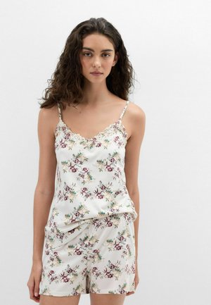 Jeune femme aux longs cheveux bouclés porte une camisole blanche ornée de dentelle florale et un short assorti sur un fond blanc uni.