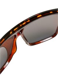 Lunettes de soleil écaille de tortue avec une monture épaisse, verres gris, finition brillante et un motif subtil le long des bords. Courbure de la monture visible.