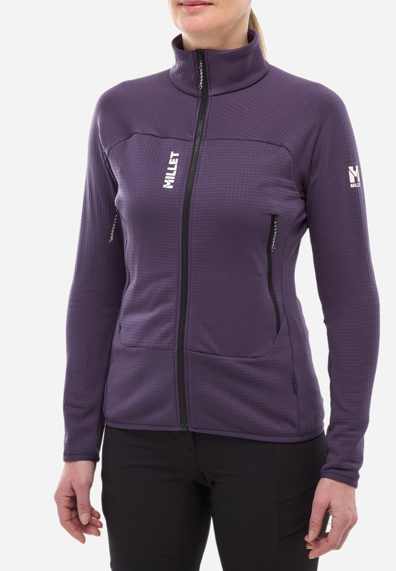 Femme portant une veste polaire zippée violette avec des logos Millet, se tenant contre un fond blanc, les mains détendues le long du corps.