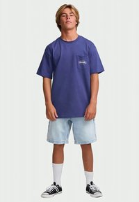 Young man standing wearing a blue t-shirt, light denim shorts, white socks, and black sneakers against a plain background.