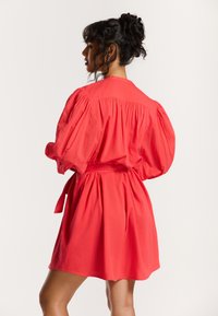 Kurzärmliges, korallenfarbiges Kleid mit tailliertem Schnitt und Puffärmeln. Hergestellt aus leichtem Stoff, mit gerafftem Rücken und Gürtel-Design.