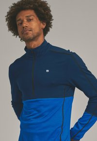 Quarter-zip pullover in dark blue and light blue fabric, featuring black stitching and a small logo on the chest. Soft texture.