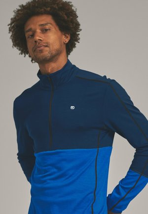 Quarter-zip pullover in dark blue and light blue fabric, featuring black stitching and a small logo on the chest. Soft texture.