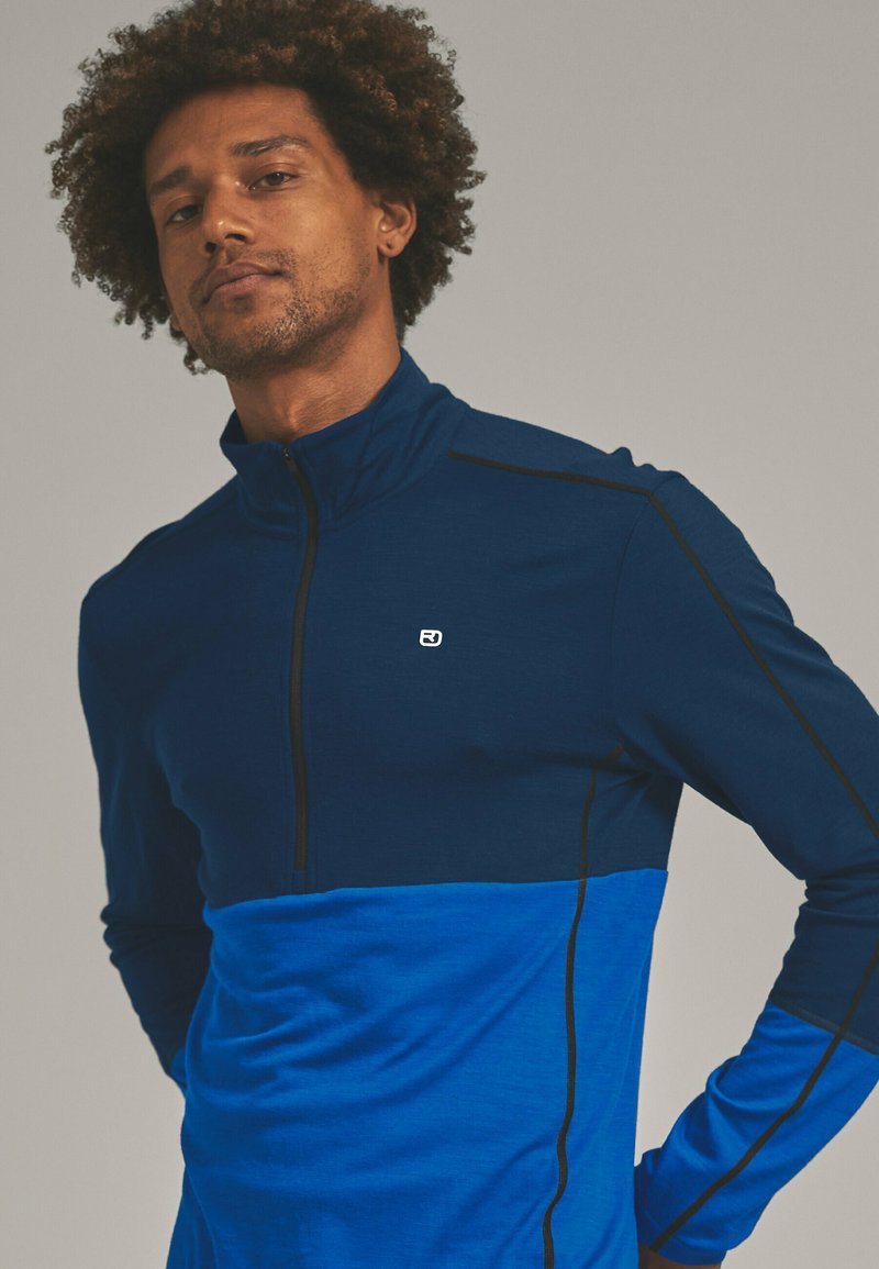 Quarter-zip pullover in dark blue and light blue fabric, featuring black stitching and a small logo on the chest. Soft texture.