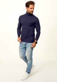 Mann mit marineblauem Rollkragenpullover, hellblauen Jeans, weißen Sneakers und einer schwarzen Smartwatch, der mit einem überkreuzten Bein nach rechts schaut.