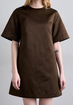 Mujer que lleva un vestido sencillo de manga corta y color marrón, con un tatuaje de libélula visible en la parte interna de su antebrazo izquierdo.