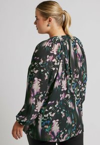 Blusa floreale nera con motivi verdi, viola e bianchi; maniche lunghe, scollo rotondo, tessuto leggero e dettaglio plissettato sul retro.