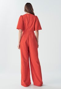 Mujer con cabello largo castaño que lleva un mono naranja brillante de pierna ancha y mangas cortas, de pie de espaldas en un fondo sencillo.