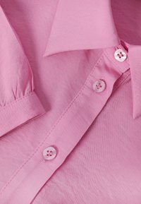 Nærbillede af en pink skjorte med knapper, der viser krave, manchet, knapper og syningsdetaljer på glat stof.