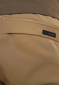 Beige broek met een gladde textuur en een bruine tailleband. Heeft een rechthoekige zak met een zwart label met de tekst "CHASIN'."