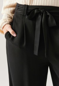 Pantalon noir taille haute avec une large ceinture, doté d'une ceinture en satin à nouer, de plis et de poches latérales, fabriqué à partir d'un tissu lisse.