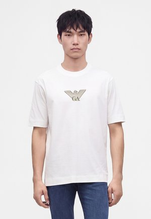 Jeune homme aux cheveux noirs courts portant un t-shirt blanc avec un logo d'aigle doré et un jean bleu, debout devant un fond uni.