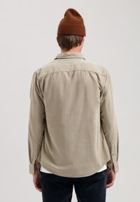 Camicia beige chiaro con una consistenza morbida, maniche a polsino con bottoni e orlo curvo. Indossata con un berretto di maglia marrone.